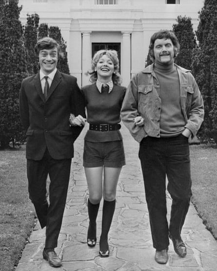 Black-and-white image of Richard O’Callaghan, Jacki Piper and Kenneth Cope walking arm in arm down a path outside the old Pinewood Studios building