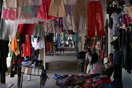 A covered market with rows of colorful clothing hanging from the ceiling, and people seated on stools.