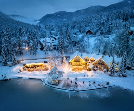 Aerial view of illuminated houses by a lake in snow with forest and mountains behind