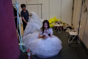 A model crouches looking at her phone while another person adjusts her white dress