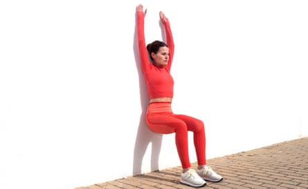 A woman does a squat against a wall with her arms raised above her head.