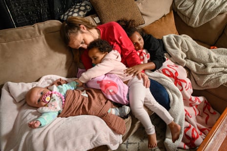 A mother and three young daughters cuddle on the couch at home.