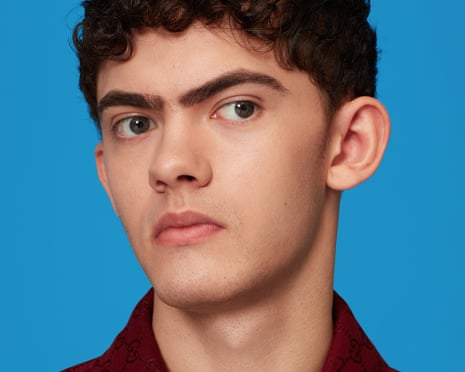 A portrait of Joe Locke, with short dark curly hair and wearing a dark red jacket, against a bright blue background