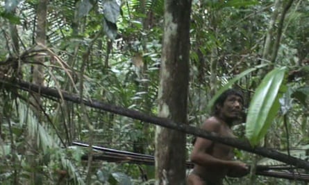 A picture of a nearly naked man carrying what look like spears as he walks though dense jungle
