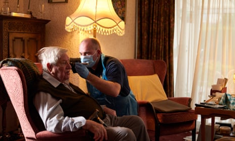 Paramedic Paul Younger, wearing a blue surgical mask, examines an older man with white hair, who is wearing a waistcoat and brown trousers, and sitting in an armchair in a home setting.