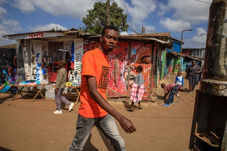 A young man in a bright orange T-shirt walks past shacks, one of which is covered in a mural