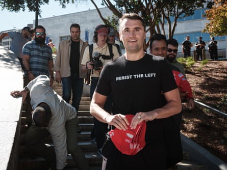 man wearing black shirt holds stack of red hats outside in front of stairs with other people behind him