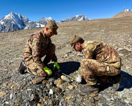 Two soldiers crouched over a small bomb with tape measure with mountain peaks in the background