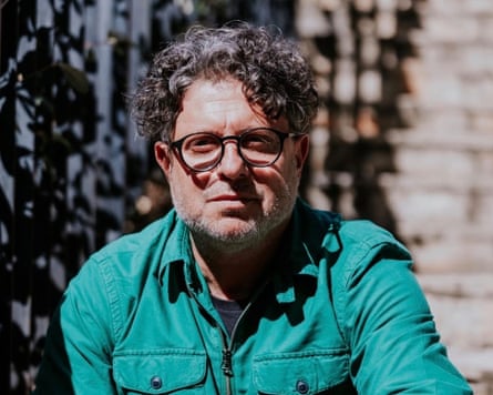 A head and shoulders shot of Isaacs, wearing glasses and a green jacket over a dark T-shirt