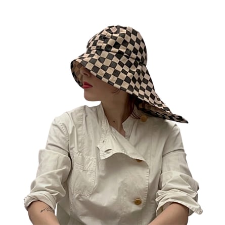 A woman wears a wide-brimmed checkered rain hat covering her eyes, over a beige coat