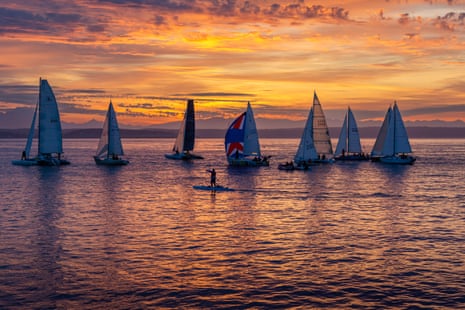 About 8 sailing yachts, and a standup paddleboard, clustered together against a backdrop of a beautiful sunrise.