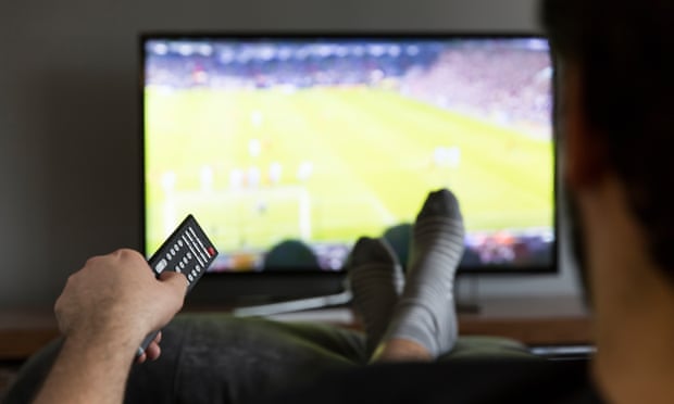 A man is watching a soccer game on television, pointing the remote at the screen, with his feet up on a cushion
