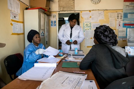 An African woman at a desk listens to a medic in protective clothing explain something while holding a sheet of paper as another woman in a white coat stands nearby