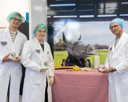Three employees in lab coats and hairnets stand by a vegan cheese display