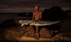A man sits at the ocean front at dusk illuminated by a yellow light, holding a surfboard on his lap