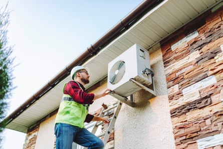 Technician in uniform repairing heat pump that is fixed to the outside wall of a house near the eaves