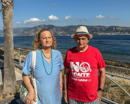Mariella Valbruzzi and Daniele Ialacqua, founders of the No Ponte Capo Peloro stand by the seafront overlooking the Strait of Messina, with mountains and coastline visible across the water.