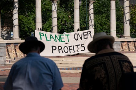 two people sitting with a banner that says “Planet Over Profits” in the background