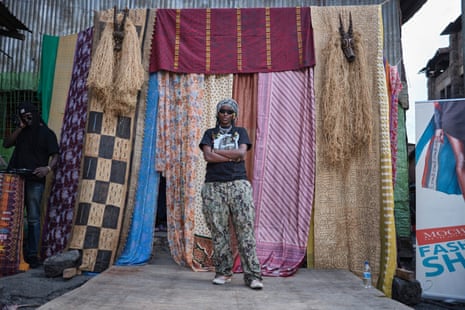A woman stands with her arms folded in front of the entrance to a catwalk hung with bright secondhand cloth.