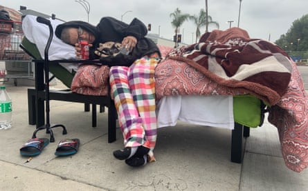 An elderly woman lying on a bed on on the road. The bed is piled with a few blankets and a walking stick stands near the head of the bed