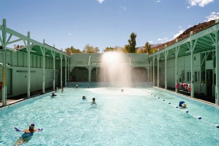 A large, rectangular, aqua pool surrounded by wooden trellises, with bathers beneath a sunny sky.