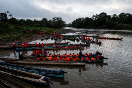 People wearing orange life jackets sit in long canoes on a riverbank