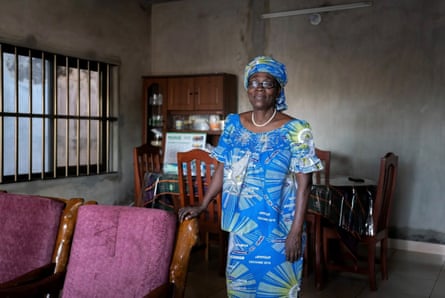 An older African woman in a matching dress and headdress poses in the living room of a house