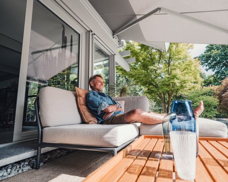 A man relaxes on a patio sofa under an awning in a sunlit summer garden