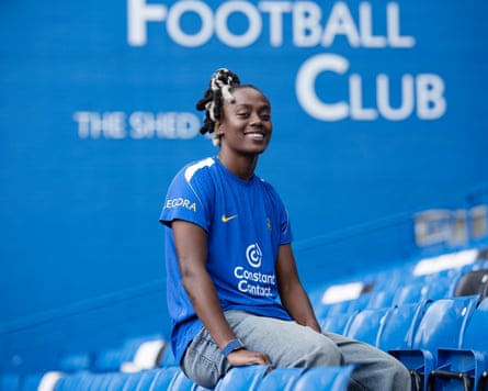 The Chelsea forward Melvine Malard smiles while posing for a photo perched on the back of a seat in the Shed End of Chelsea's home ground, Stamford Bridge. She is wearing a Chelsea training shirt and light blue jeans.