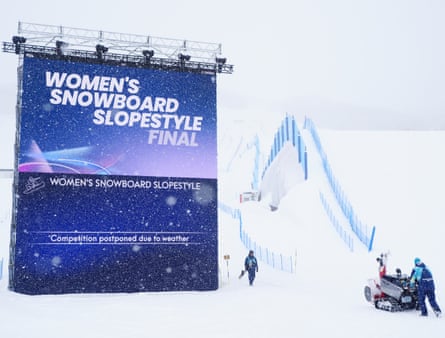 Event staff work to clear snow before the delayed women’s snowboard slopestyle final with a sign announcing the postponement at the 2026 Winter Olympics.