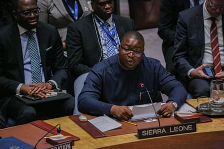 Musa Kabba sitting at a conference desk behind a microphone and sign saying Sierra Leone