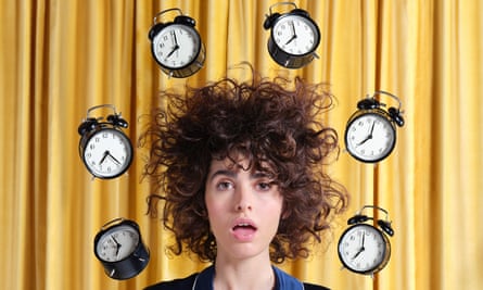 A woman with curly dishevelled hair and a ‘just woken up’ expression, surrounded by alarm clocks