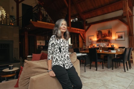 A woman wearing a black and white patterned top and black pants perches on the edge of a couch inside her home