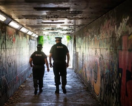A tall, strongly-built male police officer and smaller female officer are photographed from behind as they walk through a dark tunnel or underpass which has graffiti on the walls and litter on the floor.
