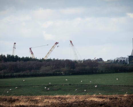 Cranes and a tall rectangular mining structure rise up behind a field of sheep.