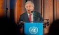 a middle-aged white man with coiffed white hair in a suit and tie speaks from behind a lectern with the light blue and white symbol of the United Nations