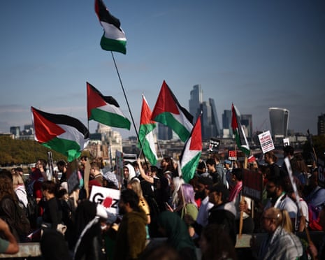 A crowd of people holding up Palestinian flags and placards about Gaza with the City of London in the background