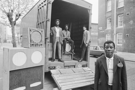 Black and white photograph of three men standing in the back of a truck parked next to a soundsys- tem. A man is also stood in front of the truck
