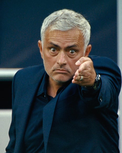 A man with grey hair with a stadium background wearing a blue suit pointing with staring eyes at someone beyond the camera