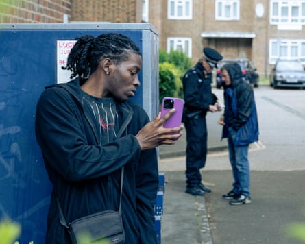 A young man films a police officer and a man, on his phone, from behind a crate