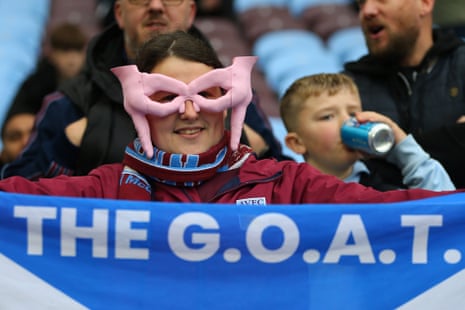 A Villa fan wears pink pig-shaped glasses, holding a blue banner reading "The G.O.A.T." for John McGinn