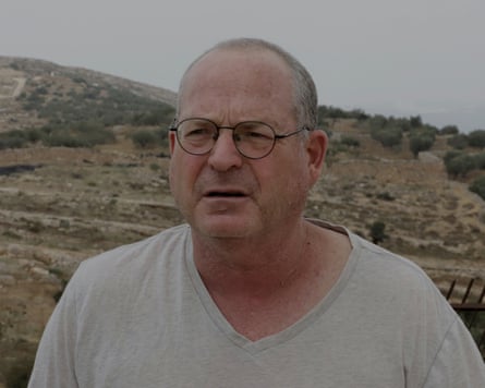 A close-up of Dror Etkes, with round glasses and a light grey V-neck T-shirt, standing outdoors in a dry, rocky landscape with scattered trees and low hills.