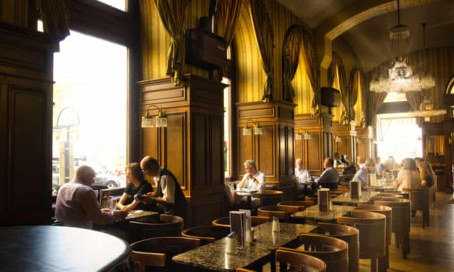 Rows of marble-topped tables with round-backed wooden chairs, huge windows with alcove seating, wood panelling, high ceilings and sun streaming in giving golden lighting to the interior of Café Schwarzenberg, Austria