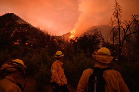 Firefighters look at a burning hillside, in orange light.
