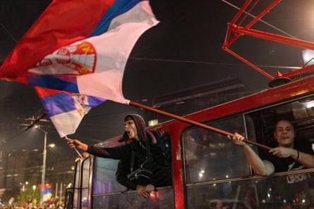 Election supporters lean from a tram at night, one waving a large Serbian flag.