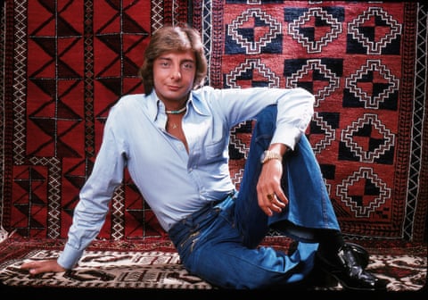 Barry Manilow in an unbuttoned pale blue shirt and navy trousers sitting on a woven rug