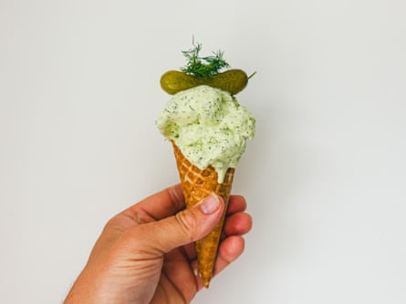 A hand holds a waffle cone topped with pale green gherkin brine ice-cream and a pickle