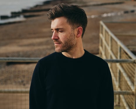 Benjamin Wood in a black T-shirt looks to the side on a beach