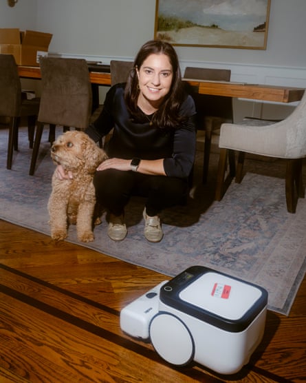 She poses in a dining room, crouched in front of a table as a robot vacuum operates. Her dog is looking away from the camera