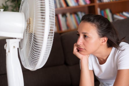 Young woman in front of electric fan; she has her hand on her chin and wears a white T-shirt. There are bookshelves in the background.